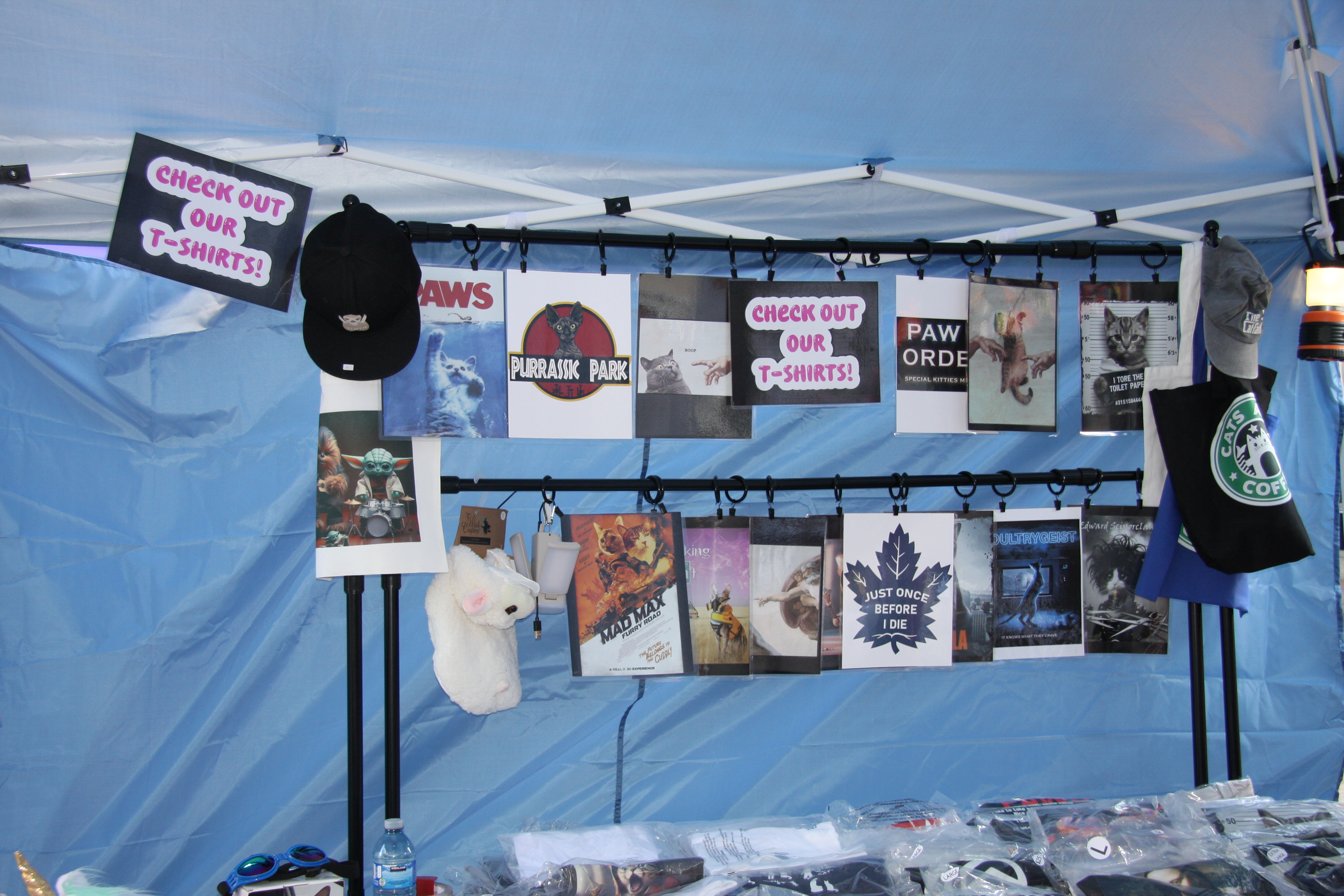 A vendor tent displaying cat-themed movie parody t-shirt designs, including "Purrassic Park," "Paws," and "Mad Max: Furry Road".