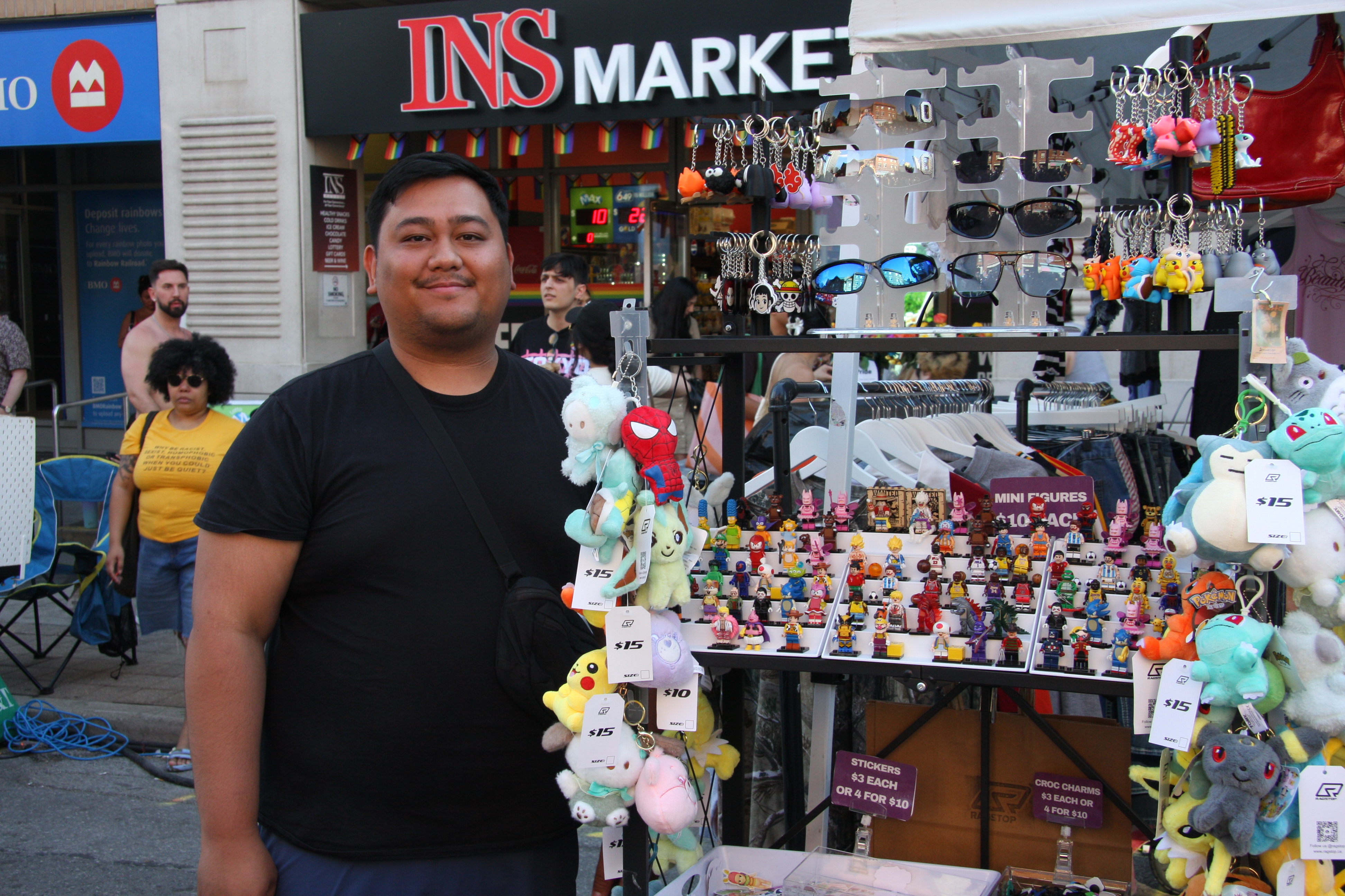 An asian man standing beside a display of collectibles, plushies, keychains, and y2k clothes.