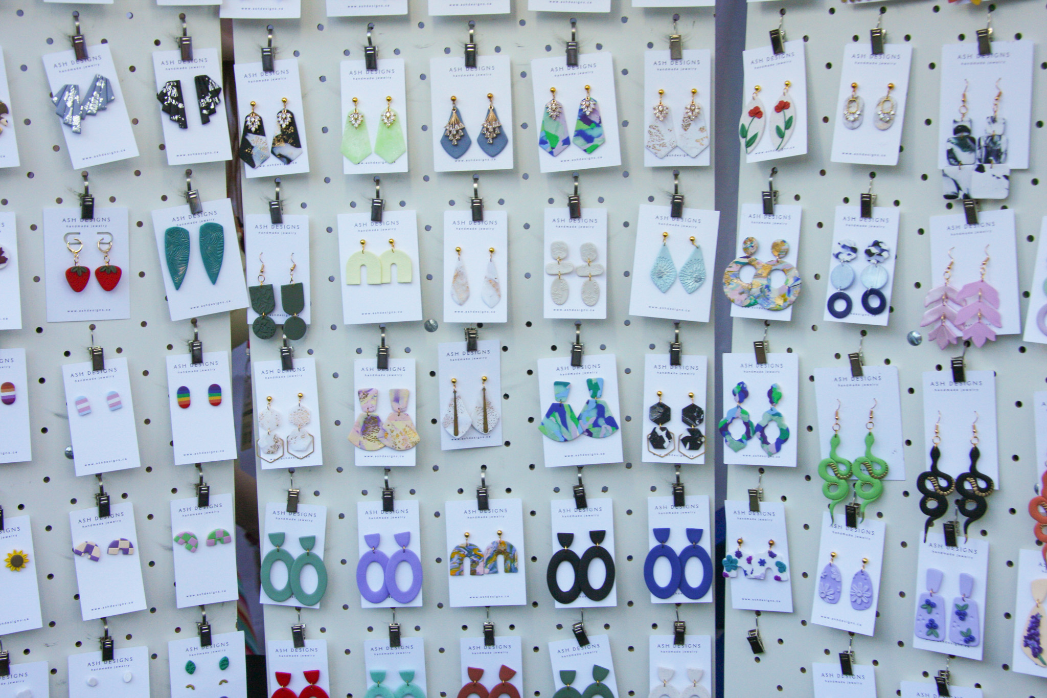 Rows of handmade polymer clay earrings by Ash Designs displayed on a market pegboard.